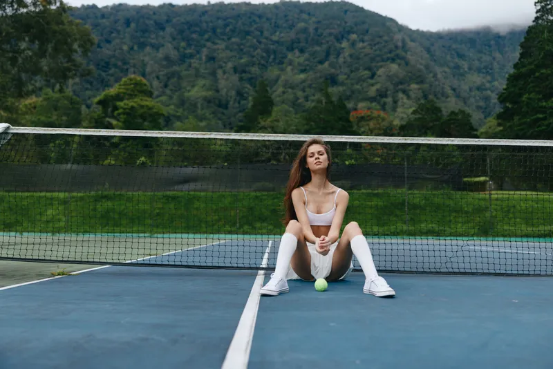 Young female athlete in sportswear, sitting on a tennis court, displaying confidence and determination against a lush green mountain background