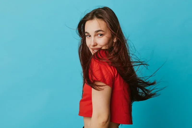 Young woman with long brown hair wearing red shirt posing and smiling against blue background. Casual portrait with natural expression and wind blowing through hair in studio setting.