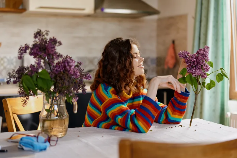 Cheerful woman indoors at a dining table, wearing a colorful striped sweater, warm sunlight streaming in, fragrant flowers in bloom, relaxed home lifestyle moment, cozy atmosphere