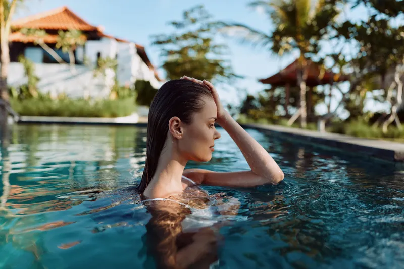 Young woman relaxing in outdoor pool, enjoying sunny day, calm water, tropical background, peaceful moment, casual lifestyle, natural light, serene atmosphere