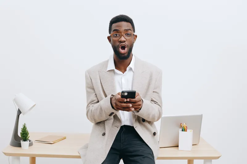 Excited young man with glasses expresses surprise while holding smartphone in office environment, casual outfit, modern workspace, white background, emphasizing emotion and technology use, lifestyle c