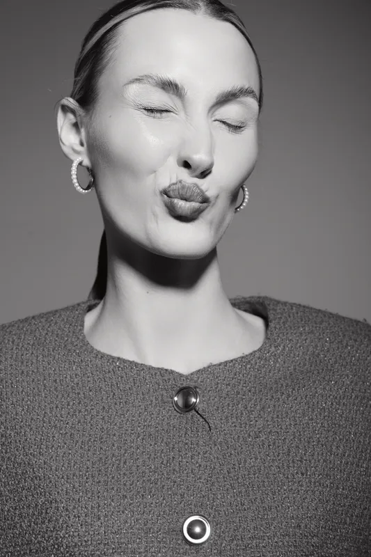 Portrait of a smiling white woman with fair skin and slicked hair, eyes closed, making a playful kiss face, wearing earrings and textured top in studio lighting.