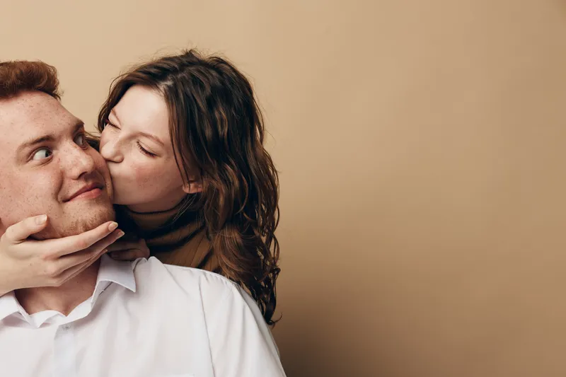 Young woman kisses smiling man on cheek while holding his face gently. Intimate moment between couple on beige background. Concept of love, affection, and relationship.