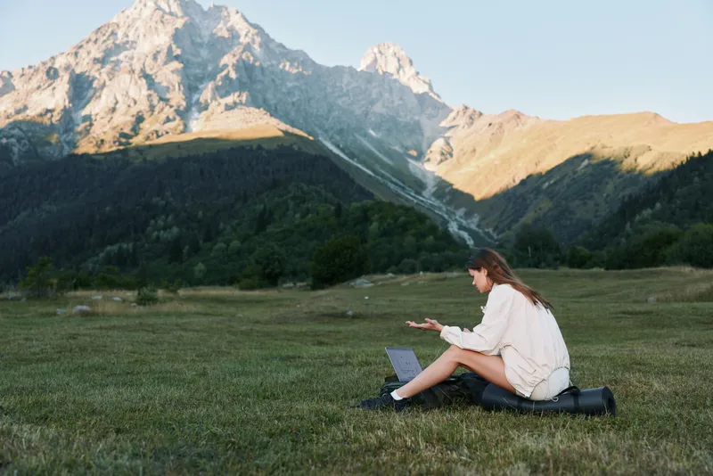A lone traveler sits on a grassy field with a laptop, immersed in work as dramatic mountains rise behind, creating a serene outdoor workspace.