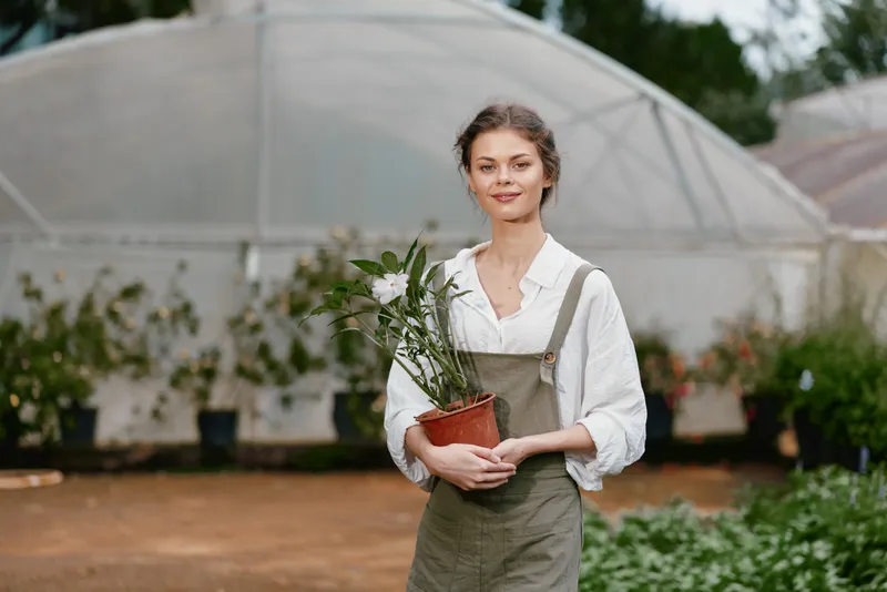 Young woman in casual overalls holding a potted plant in a greenhouse, embodying a healthy gardening lifestyle with natural beauty and joy