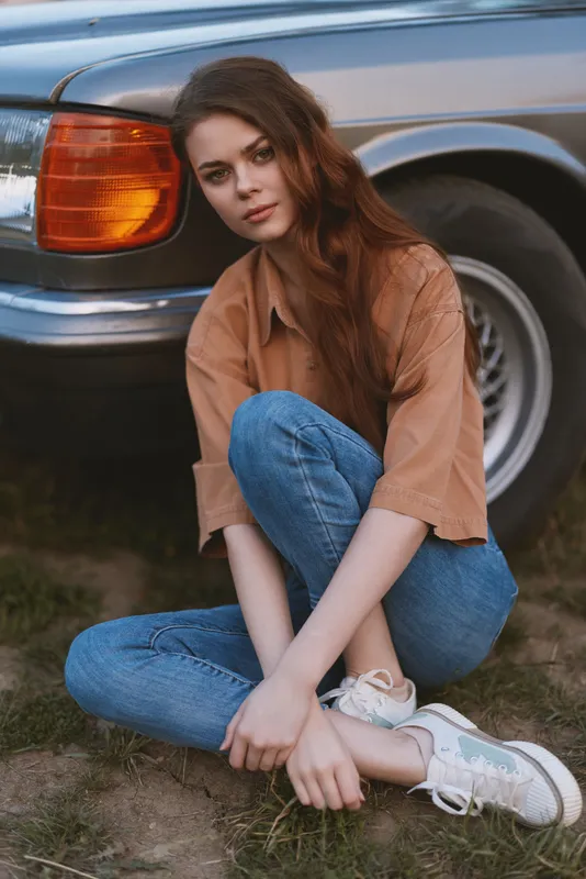 Young woman in casual summer outfit sitting by vintage car, expressing relaxed emotions, with soft natural background and warm earth tones.