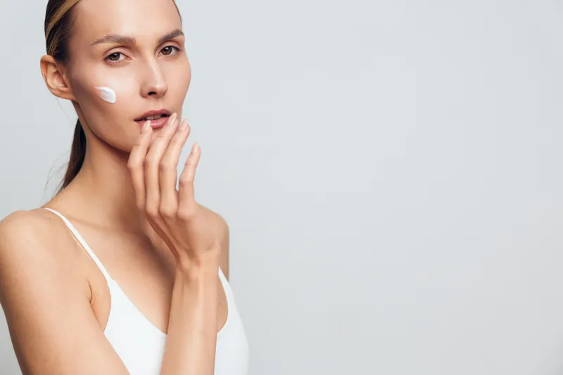 Young woman with clean skin applying face cream on cheek, wearing white tank top, posing against light gray background. Concept of skincare, beauty, hydration, and natural wellness.