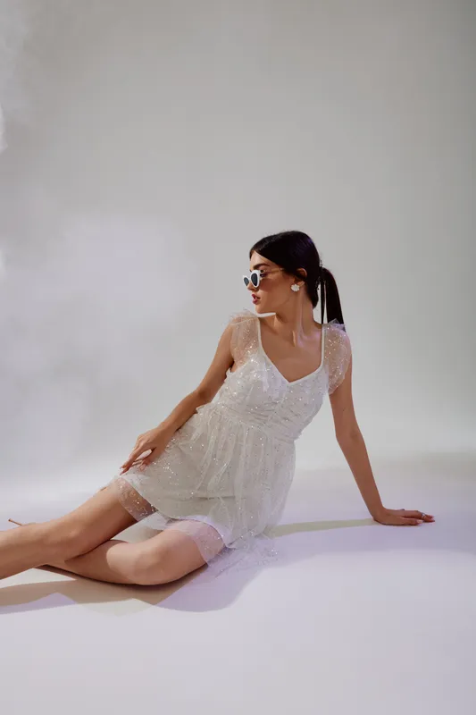 Young woman in a white lace dress and sunglasses sits on the floor against a plain background. Fashion concept highlighting beauty, style, and elegance in studio lighting.