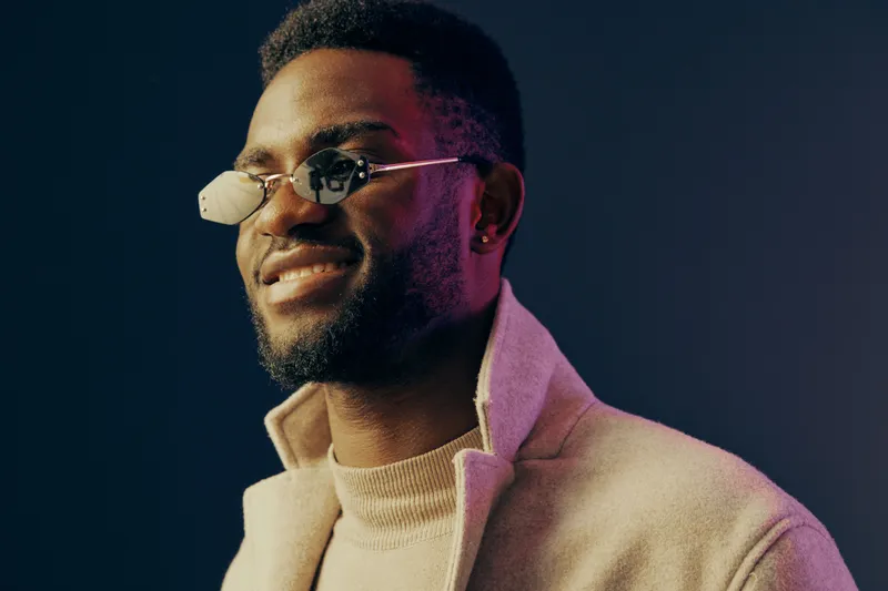Stylish young man wearing modern glasses, smiling confidently against a dark background, showcasing a contemporary fashion aesthetic with a warm color palette