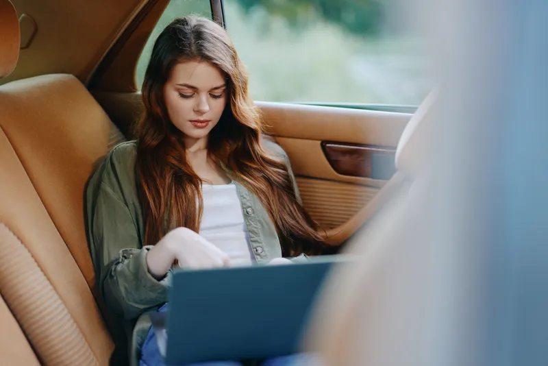 Young woman working on a laptop in a car, wearing casual clothes, with a serene expression and natural background, conveying productivity and focus.