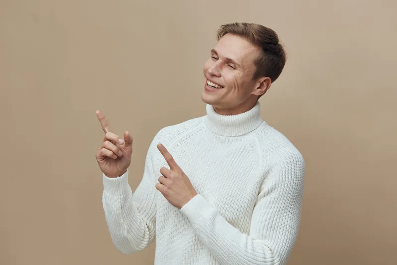 Smiling happy man in white sweater pointing upwards with both hands on beige background, casual confident expression, lifestyle concept, cheerful mood, portrait photography.