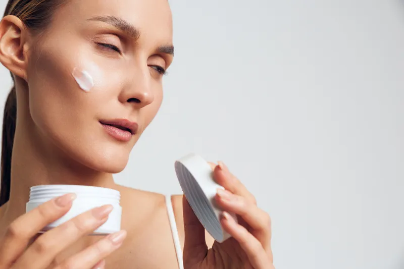 Young woman with closed eyes applies white face cream on her cheek while holding an open jar and lid in soft natural light on a plain background.