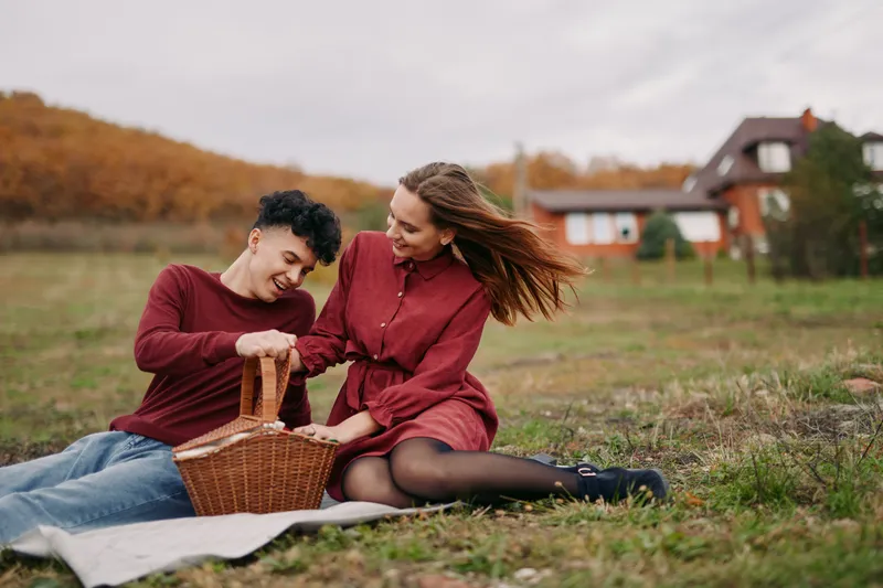 A candid outdoor picnic portrait of a couple sharing a wicker basket on a blanket in an autumn field, natural interaction and gestures that convey authenticity and quiet credibility.