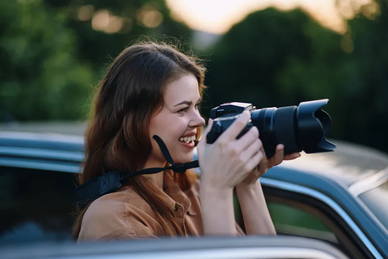 Young female photographer capturing nature with her camera, showcasing a joyful expression and outdoor scenery during golden hour.