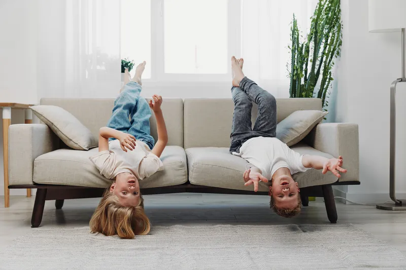 Two happy children hang upside down from a beige sofa in a bright living room with plants, enjoying playful moments and laughter. Concept of childhood fun and carefree home environment.