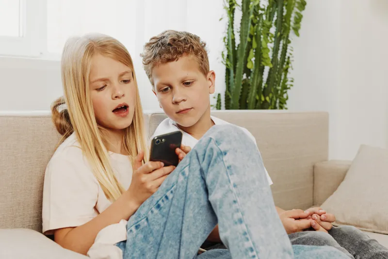 Two children sitting on a couch looking at a smartphone together with focused expressions in a bright living room. Concept of technology, childhood, and shared curiosity.