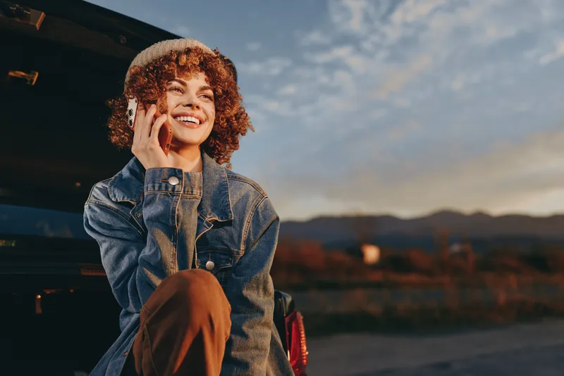 Woman lifestyle moment, smiling woman wearing denim jacket, holding phone to ear, curly hair, warm sunset sky, outdoors in evening air, relaxed casual travel mood, city background blur