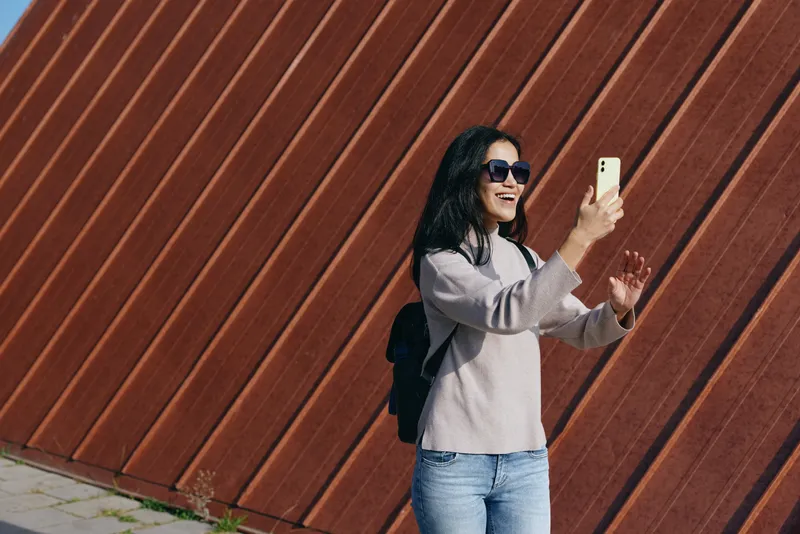 Selfie of a woman with sunglasses and backpack filming herself with a smartphone outdoors, against a diagonal wooden wall, casual style and confident smile, urban vibe and travel mood.