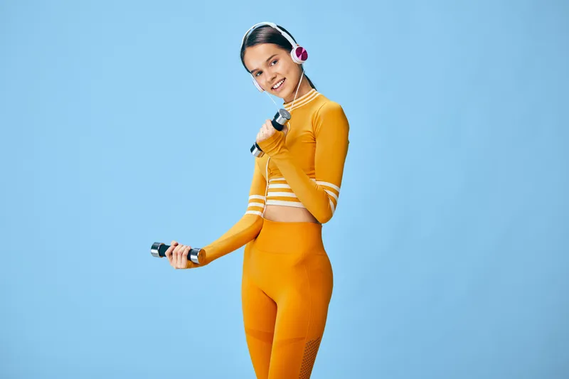 Woman in bright orange fitness outfit poses with a dumbbell and headphones against a blue studio backdrop, showcasing athletic style, focus, and energetic movement.