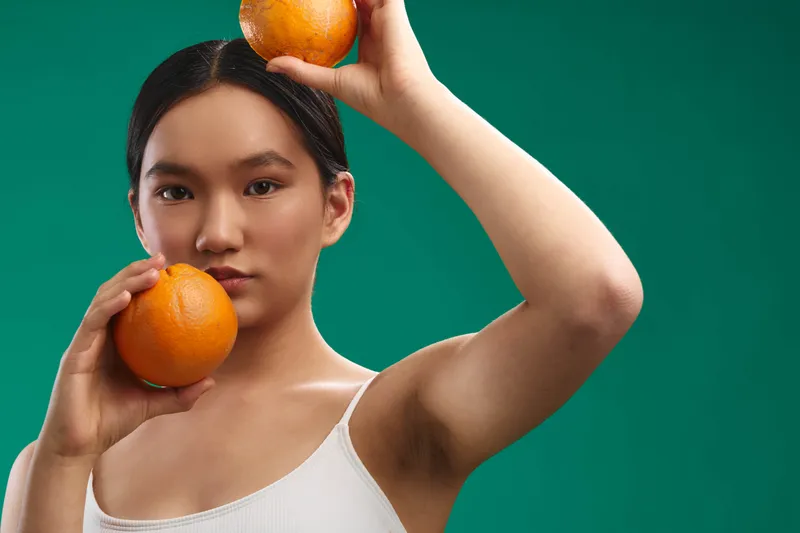 asian woman holding oranges in a bright studio, calm and confident expression, conveying a fresh, vibrant lifestyle moment with a modern, creative everyday mood.