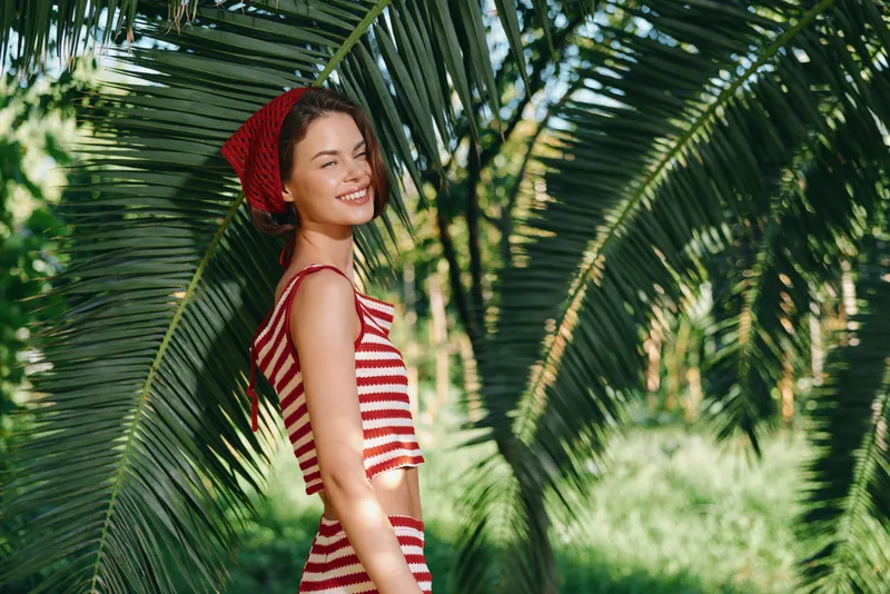 A cheerful young woman in a red striped crop top stands outdoors among palm fronds, smiling warmly, capturing a bright, relaxed tropical moment.