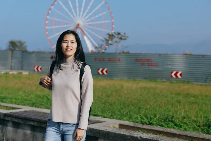 Woman in a light sweater and jeans stands in a park with a large ferris wheel in the background, casual outdoor scene, daytime travel mood, relaxed confident stance and sunny atmosphere, bright open