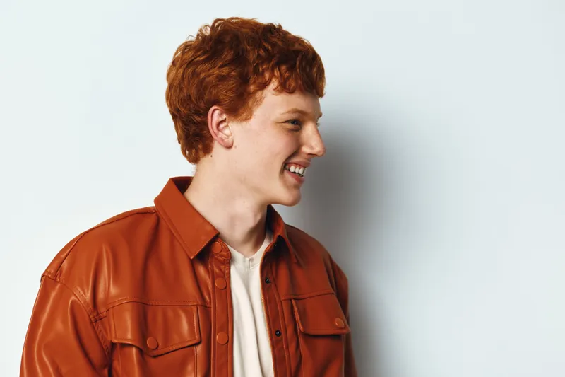 Young man with red hair wearing brown leather jacket smiling looking to the side on light background. Casual style portrait of cheerful male adult in studio with curly hair and white shirt.
