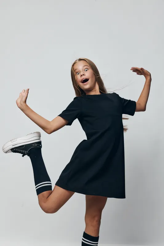 Girl jumping and posing with energetic facial expression wearing black dress, striped socks and white sneakers against neutral background. Youth, fun, movement and casual style.