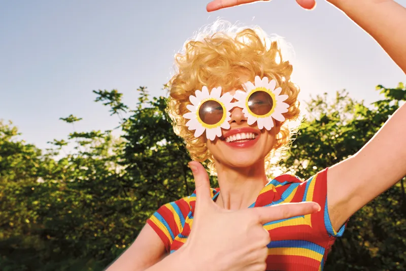 Smiling woman with flower sunglasses outdoors. Retro film color and sunny background convey vibrant lifestyle, playful mood, and carefree summer vibe.