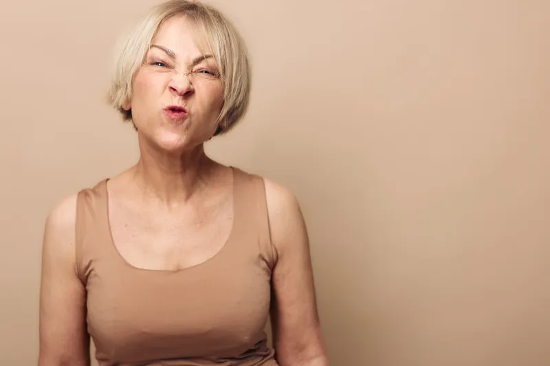 Mature woman with short blonde hair makes a funny grimace with scrunched nose and pursed lips, wearing a beige tank top against a neutral background. Concept of humor, expression, and emotion.