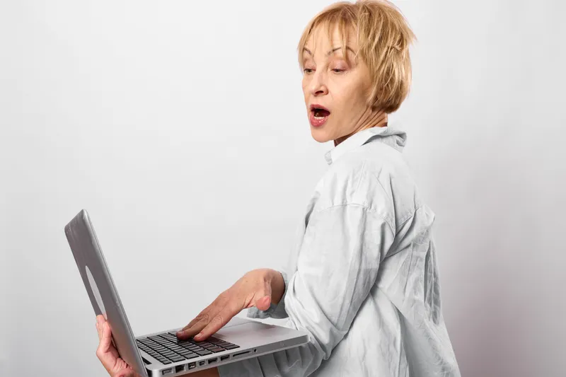 Woman holds an open laptop on a white background, looks surprised and shocked, mouth open with panic, office worker posture, technology stress moment instant response