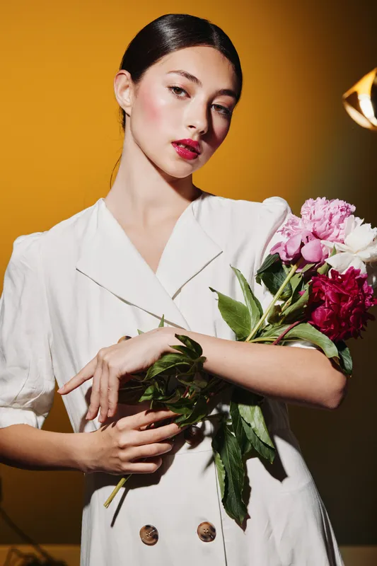 Elegant woman in white dress holding colorful flowers. Creative fashion shot with vibrant makeup on a warm orange background. Concept for beauty, style, and natural elegance.
