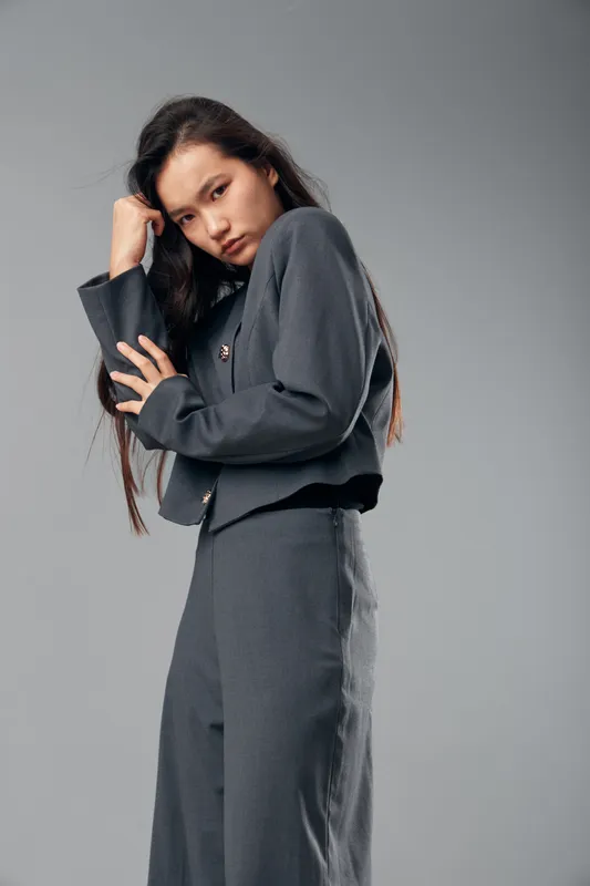 Woman posing in grey formal outfit standing against plain grey background with crossed arms and thoughtful expression, fashion style portrait with long dark hair and minimal makeup.