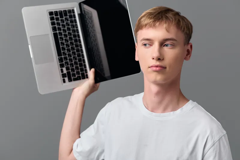 Laptop held near the head by a young man, illustrating tech exploration, learning and focus in a casual studio setting with a modern device, gadget usage and a clear education mindset