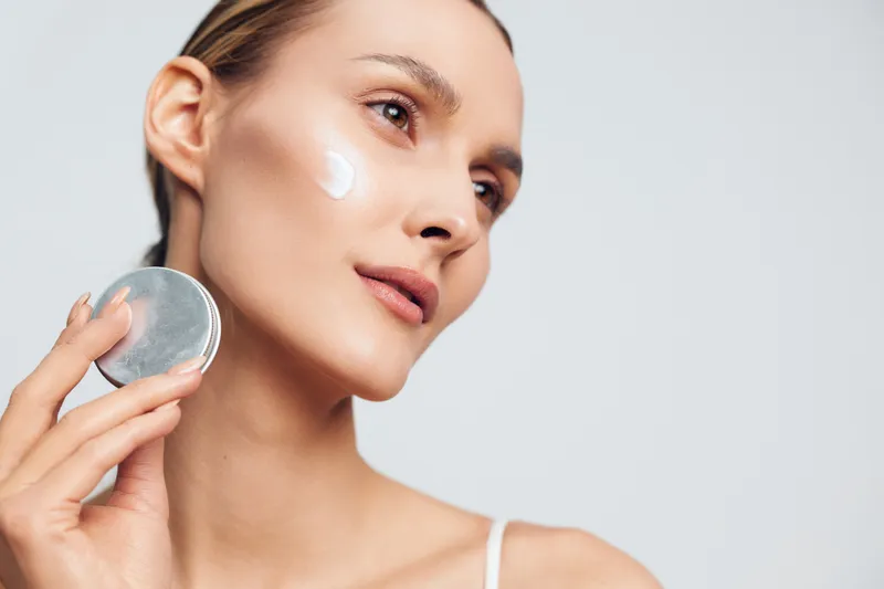 Young woman with blonde hair applies moisturizing cream on her cheek while holding a cosmetic jar against a light background with copy space.