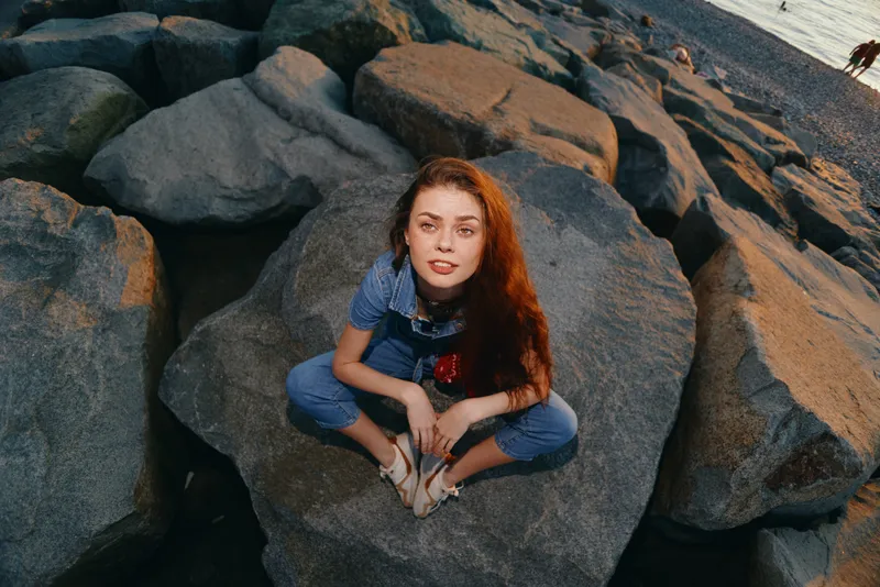 lifestyle woman in modern boho-western denim street style sitting on rocks with natural film color tones, relaxed summer atmosphere by the sea shore