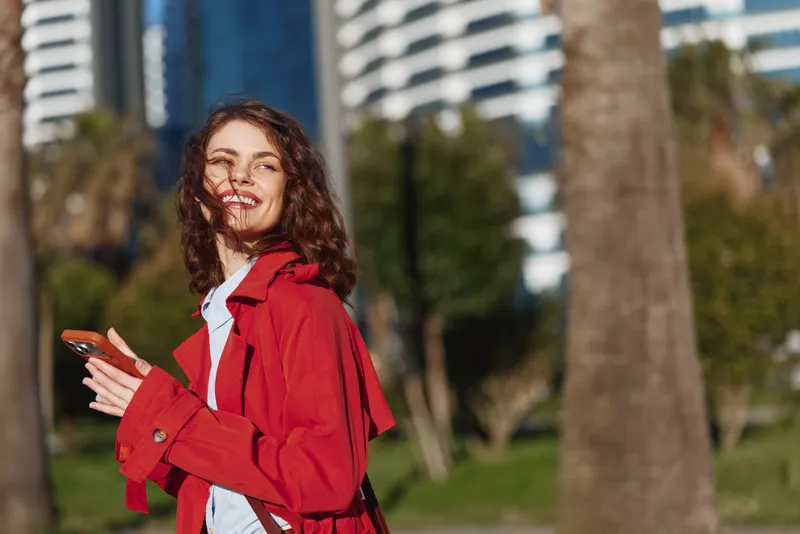 Young woman with curly brown hair smiles joyfully while holding a smartphone outdoors in a sunny urban park, wearing a red jacket and white shirt during a relaxed moment.