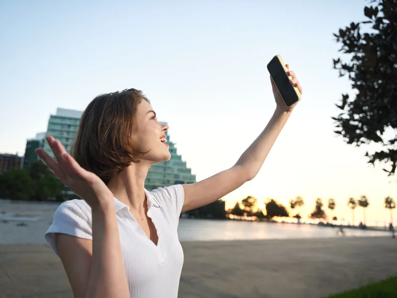 A cheerful woman in casual attire takes a selfie outdoors with a smartphone, smiling at the camera as sunlight frames her portrait in a bright, carefree moment.