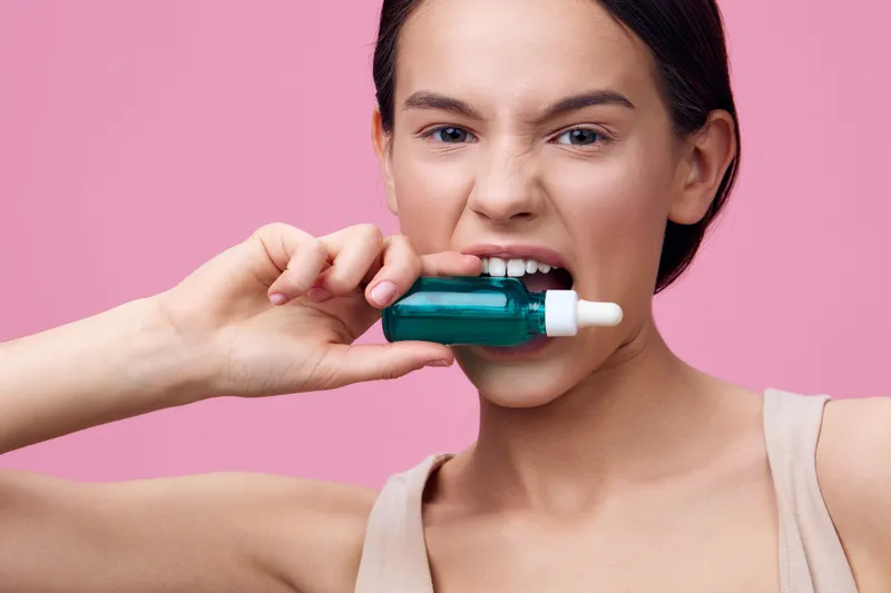 Young woman showing a skincare serum bottle, portraying confidence and fun against a pink background, ideal for beauty and skincare promotions