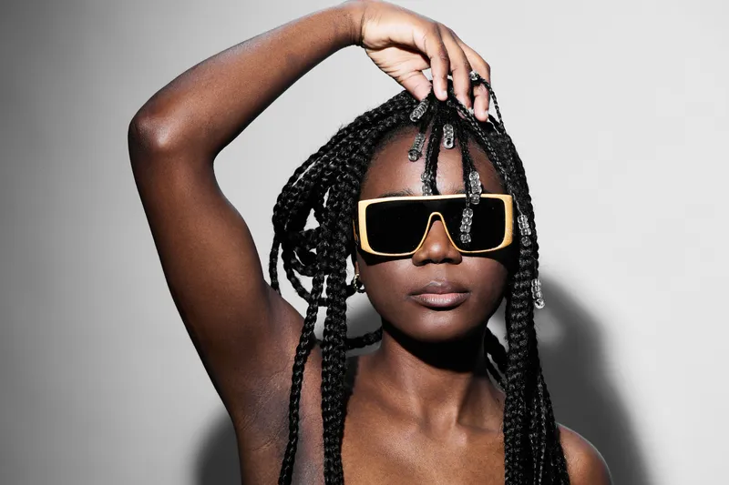 Confident young woman with braided hair showcasing stylish oversized sunglasses against a minimalistic backdrop, embodying modern fashion trends