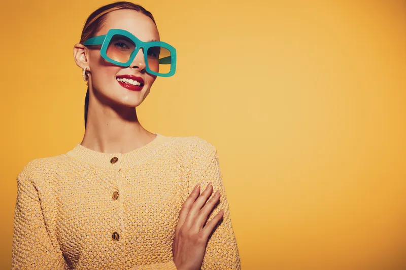 Stylish woman with blue oversized sunglasses smiles confidently wearing a textured yellow sweater against a bright yellow background. Concept of fashion, summer, and vibrant style.