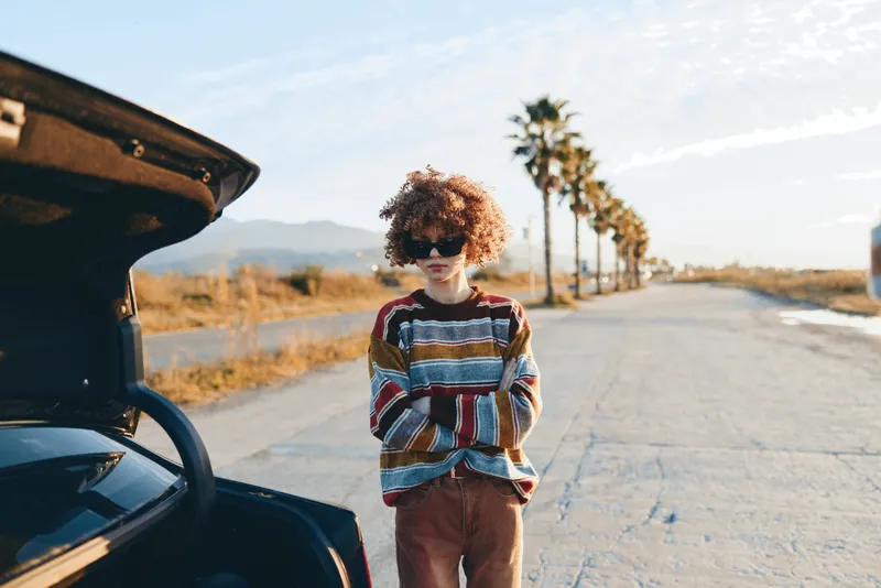 Young woman lifestyle on a sunny road, standing beside an open car hood, curly hair and sunglasses, relaxed travel mood, palm trees and dry hills in the distance, golden light