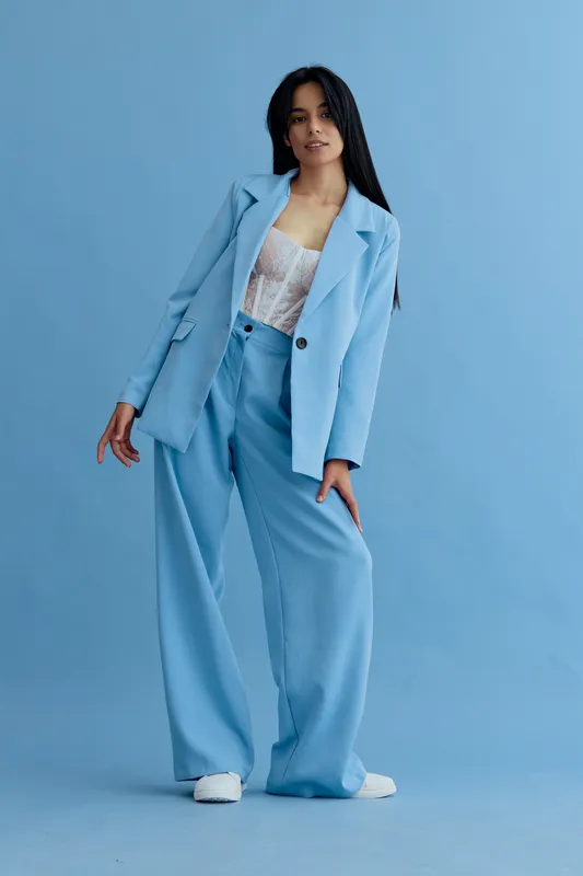 Young woman in stylish light blue suit posing confidently with casual lace top and white sneakers against a matching blue studio background, exuding modern fashion vibe.