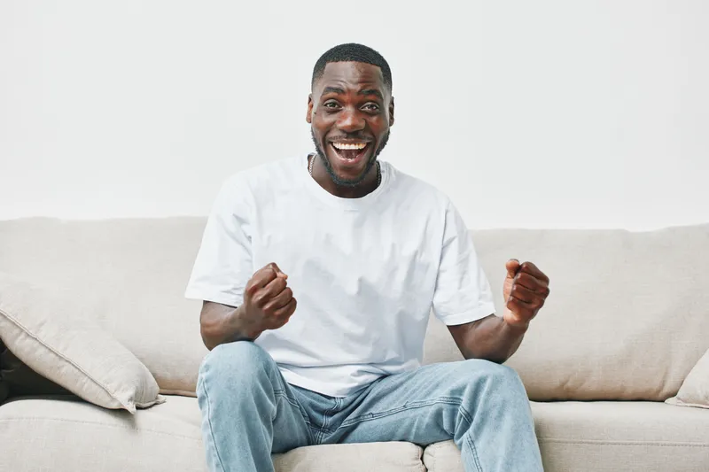 Happy young African man sitting on a couch, expressing joy and excitement in a bright, minimalistic living room setting