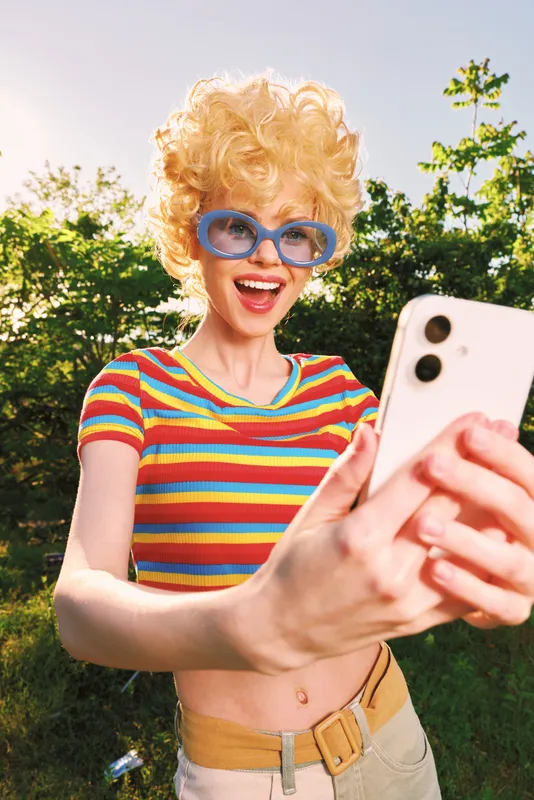 A young woman with curly blonde hair and oversized blue sunglasses snaps a playful selfie in a sunny park, wearing a striped crop top for a retro vibe and bold color.