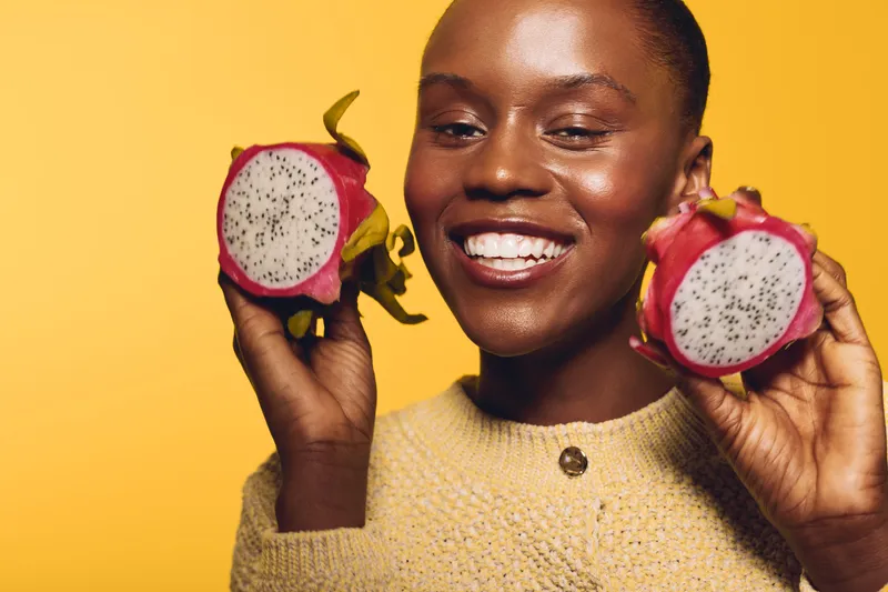 Smiling dark skinned African woman holds two halves of a dragon fruit near face. Studio portrait on yellow background with bright mood. Concept of healthy lifestyle, exotic fruit and happiness.