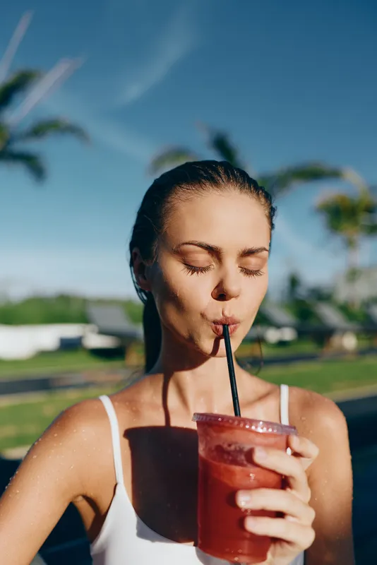 A woman enjoys a bright poolside beverage under a sunny sky, sipping through a straw with eyes closed. Casual summer moment capturing relaxation and refreshment.