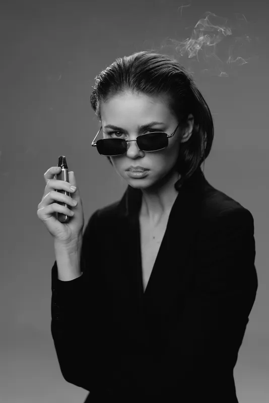 Black and white portrait of a serious woman in sunglasses holding a vape with smoke around her.