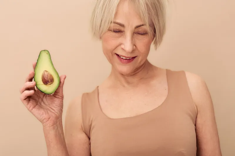 Smiling mature woman with short blonde hair holds half of avocado near face on beige background. Concept of healthy eating, natural beauty, wellness, and fresh organic food lifestyle.