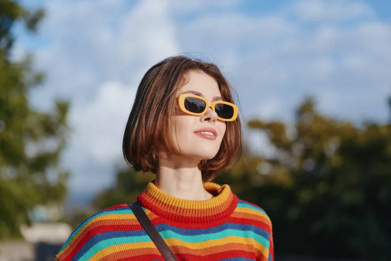 Woman young adult in colorful striped sweater and yellow sunglasses smiling outdoors in a sunlit urban park; short bob haircut, casual outfit, looking up for travel lifestyle and fashion.